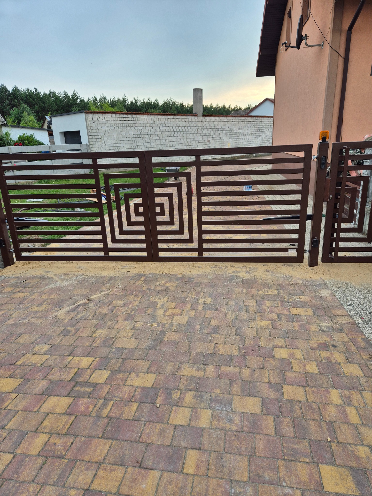Nowoczesna brama wjazdowa w kolorze brązowym, z geometrycznym wzorem kwadratów. Widoczny automat do bramy i kostka brukowa w odcieniach brązu i żółci.