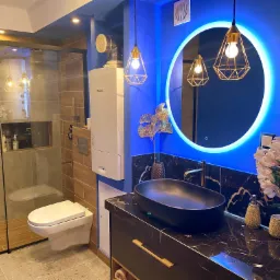 Nowoczesna łazienka z czarną umywalką na blacie z marmuru, podświetlanym okrągłym lustrem i kabiną prysznicową ze szklanymi drzwiami, oświetlona wiszącymi lampami z geometrycznymi kloszami.