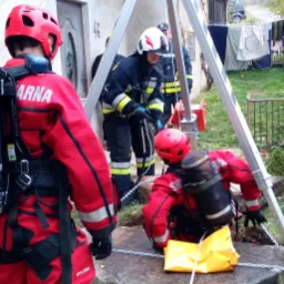 Strażacy w czerwonych kombinezonach zabezpieczają studnię za pomocą trójnogu ratowniczego i liny.