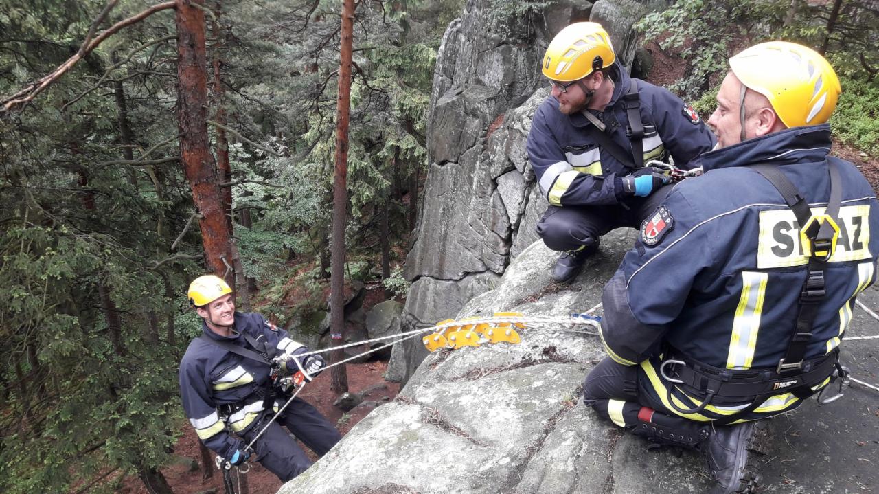 Trzech mężczyzn w kaskach i kombinezonach na skalistej powierzchni, zabezpieczonych linami, prawdopodobnie podczas treningu lub akcji ratunkowej w trudnym terenie leśnym.