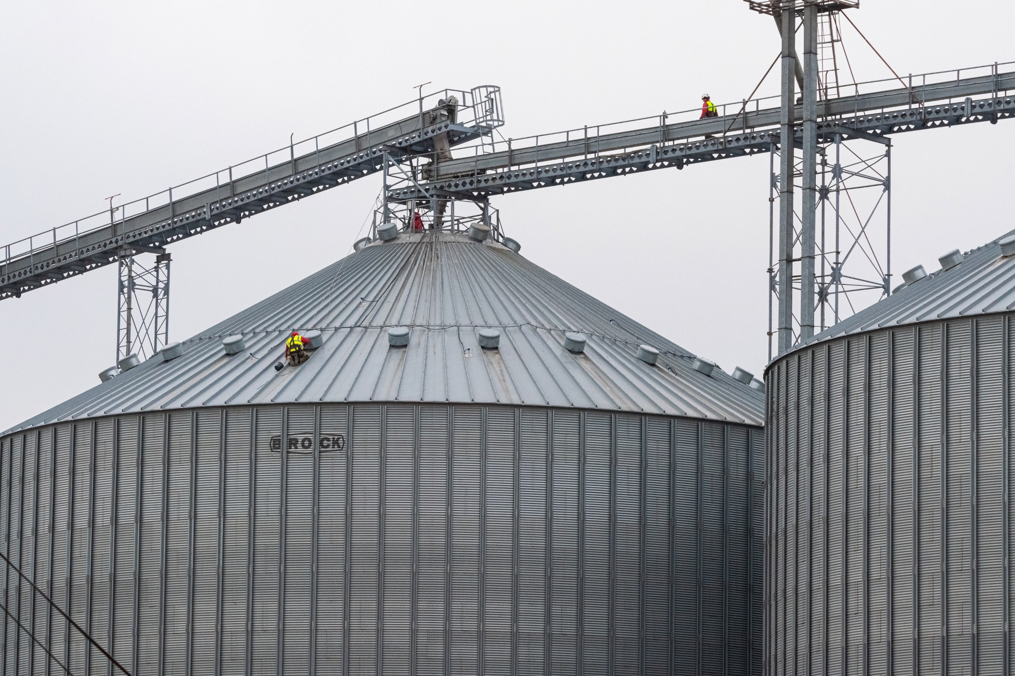 Pracownik w żółto-czerwonej kamizelce na szczycie metalowego silosu zbożowego z napisem 'ROCK', drugi pracownik na metalowej kładce nad silosem, pochmurne niebo w tle.