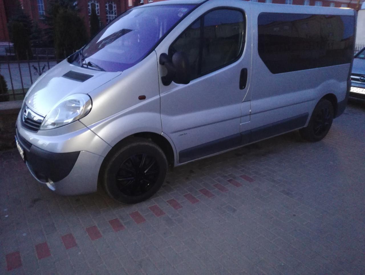 Srebrny Opel Vivaro z przyciemnianymi szybami i czarnymi felgami, zaparkowany na chodniku z czerwonymi akcentami.