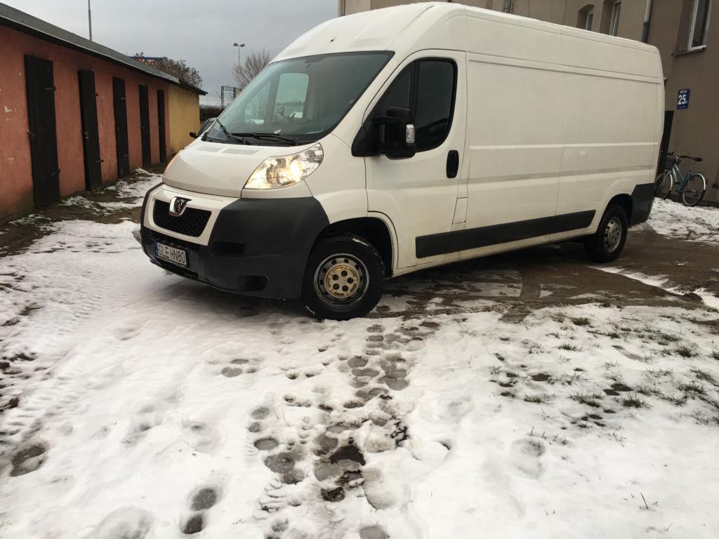 Biały furgon dostawczy marki Peugeot z włączonymi światłami, zaparkowany na zaśnieżonym placu przed budynkiem z czerwonymi drzwiami garażowymi.