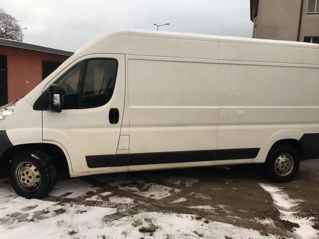 Biały bus dostawczy na tle budynku w pochmurny dzień, z brudnymi oponami i śladami kół na śniegu.