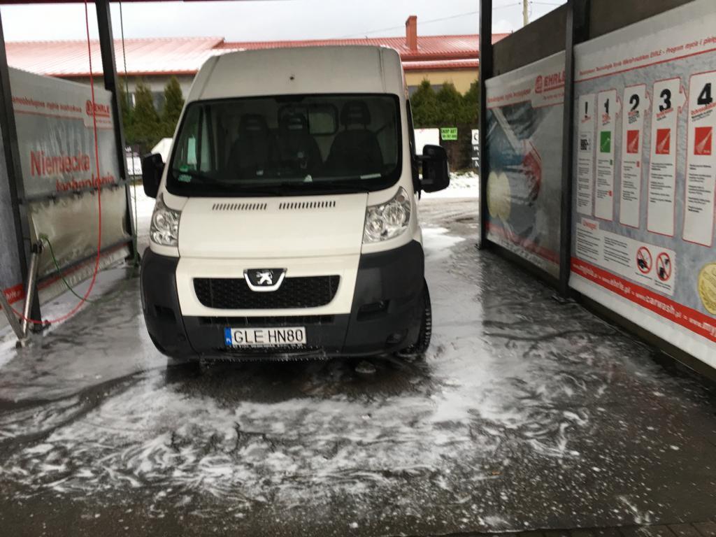 Biały bus marki Peugeot, z widoczną tablicą rejestracyjną, podczas mycia w myjni samochodowej, otoczony pianą.