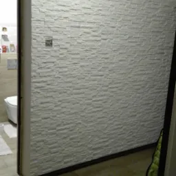 Ściana wykończona dekoracyjną, białą okładziną imitującą kamień, widoczna z perspektywy wejścia do łazienki z toaletą i akcesoriami. Na ścianie znajduje się gniazdko elektryczne.