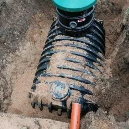 Czarny, żebrowany zbiornik z zieloną pokrywą częściowo zasypany piaskiem w wykopie, pomarańczowa rura kanalizacyjna w ziemi.