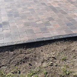 Nowa nawierzchnia z kostki brukowej w odcieniach szarości i brązu, obramowana ciemnymi obrzeżami, przylegająca do przygotowanego gruntu z fragmentami trawy.