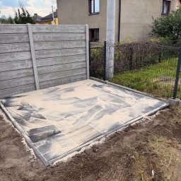 Kamil Gaweł - Ułożona, kwadratowa powierzchnia z szarych płyt, zasypana piaskiem, otoczona ziemią. W tle betonowy płot i budynek mieszkalny. Widok z góry, w słoneczny dzień.
