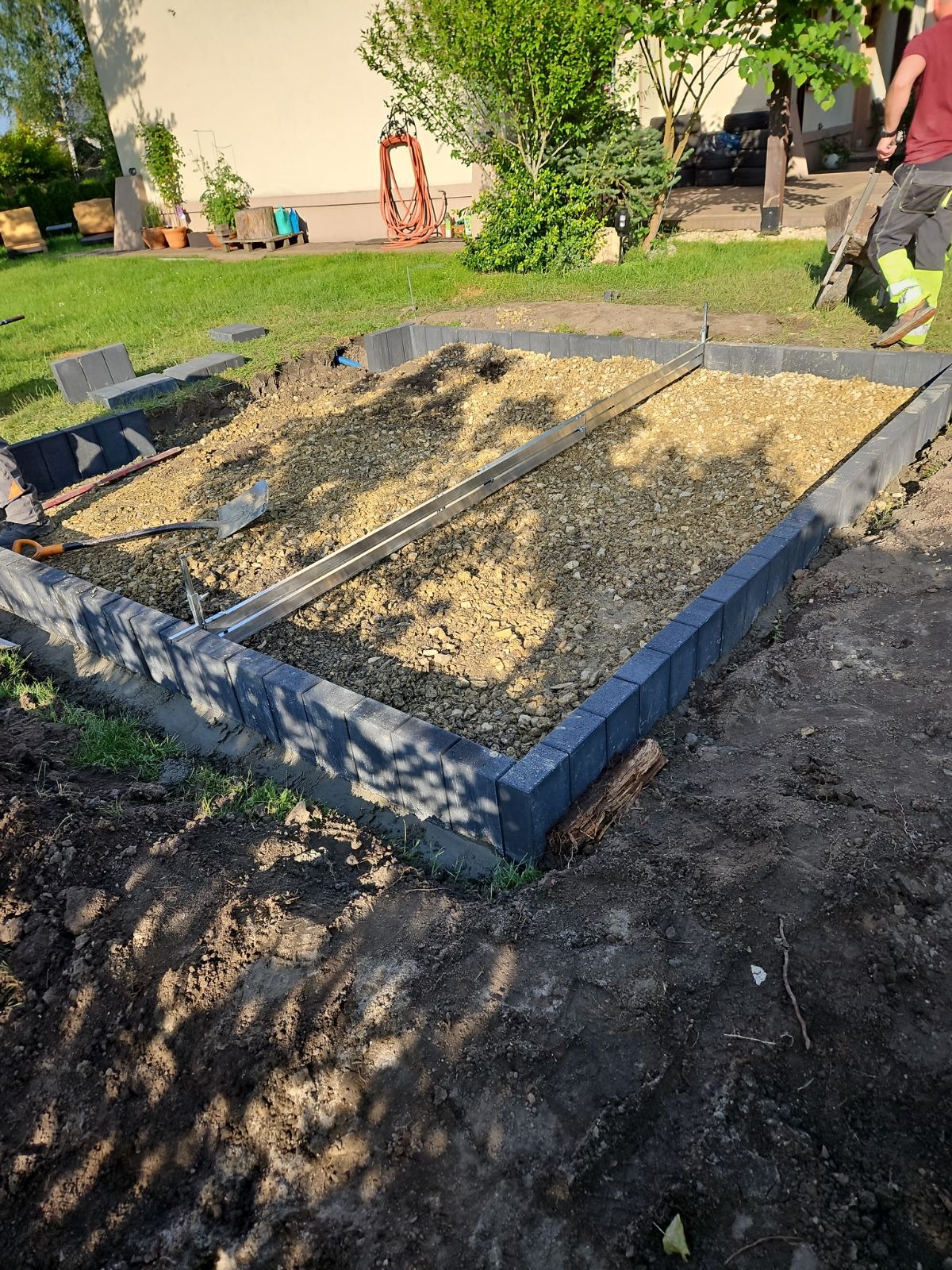Krawężnik z szarej kostki obramowujący teren wypełniony żwirem, z poziomnicą i narzędziami w środku. Widoczne trawnik i budynek w tle. Prace ziemne w ogrodzie.