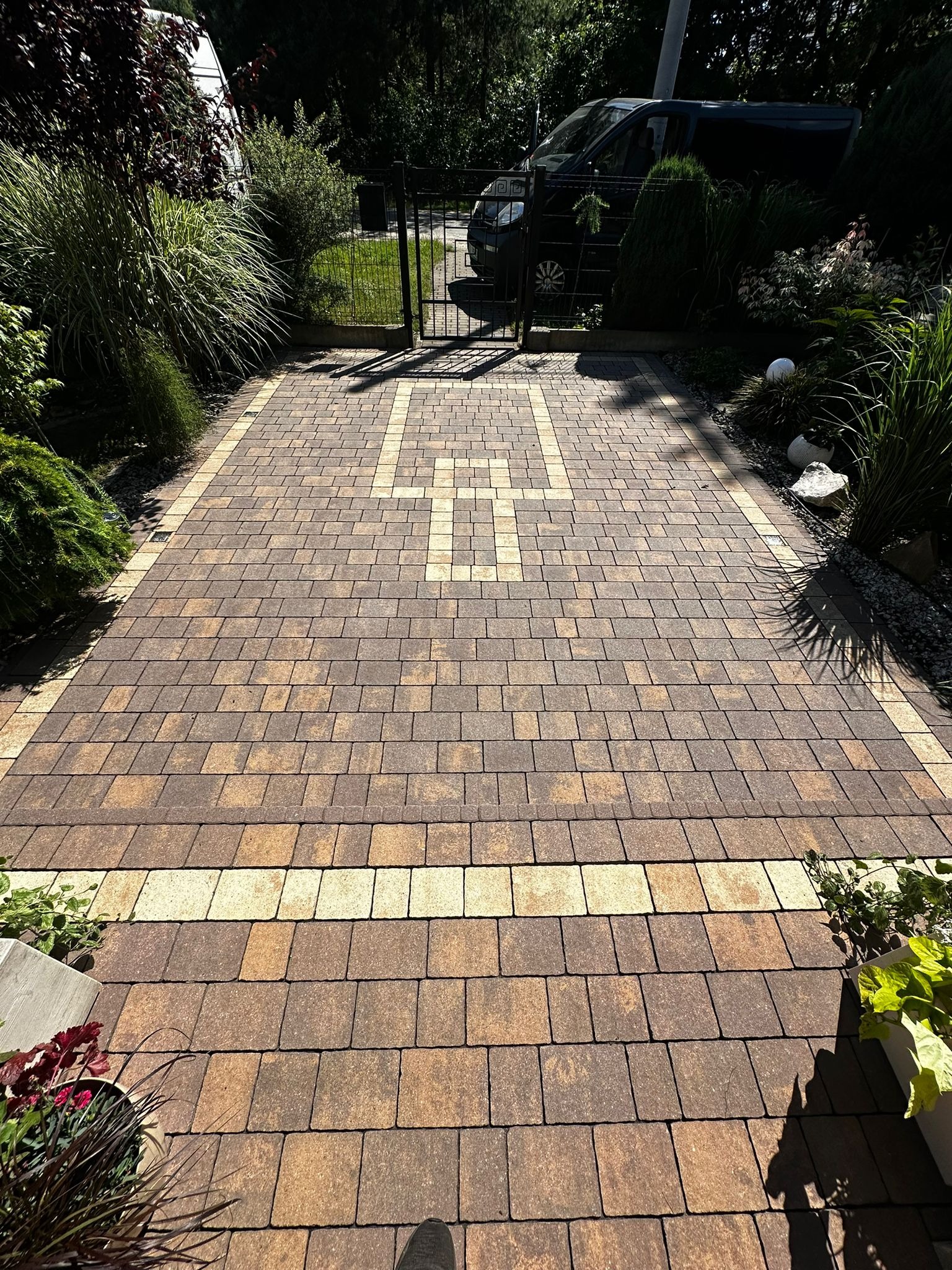 Ułożona kostka brukowa w odcieniach brązu i beżu, tworząca wzór z jasnymi elementami. Widok na bramę i zieleń ogrodu. Częściowo widoczny but osoby fotografującej.