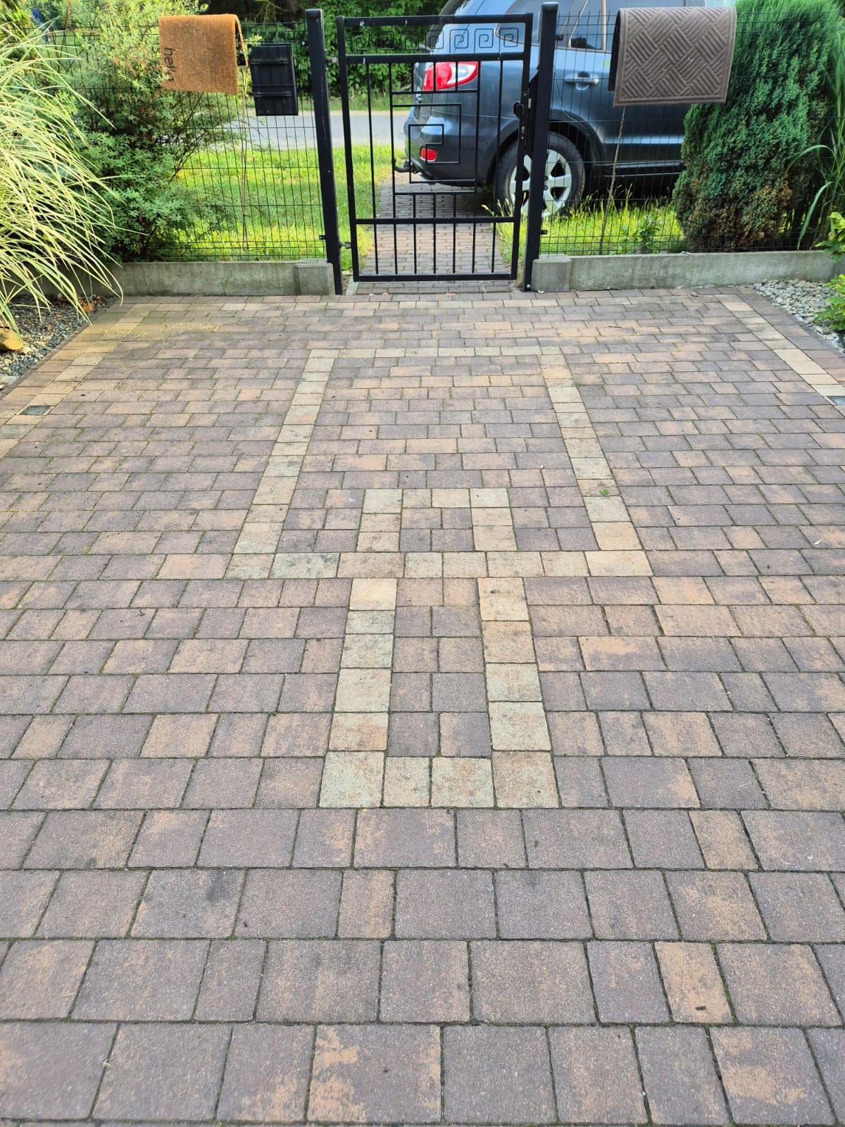 Ułożona kostka brukowa w geometryczny wzór na podjeździe, z bramką i fragmentem samochodu w tle. Widoczne wycieraczki na bramce i zadbana zieleń.