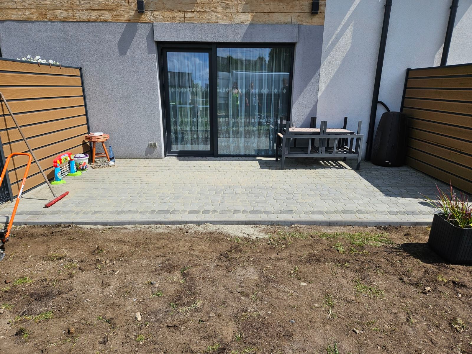 Ułożona kostka brukowa na tarasie domu jednorodzinnego, widoczne narzędzia ogrodnicze i przygotowany teren pod dalsze prace. Jasne, naturalne światło.
