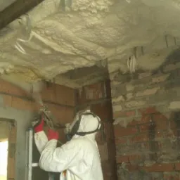 Pracownik w kombinezonie ochronnym aplikuje piankę izolacyjną na sufit poddasza za pomocą pistoletu natryskowego; widoczne drewniane belki i fragmenty muru z cegły.