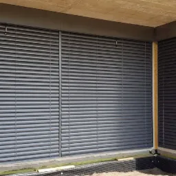 Szare poziome lamele osłaniające okna w surowym stanie budynku, widoczne cegły i brak wykończenia elewacji.