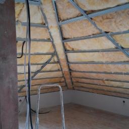 Pepo - Poddasze w trakcie prac wykończeniowych: widoczne ocieplenie wełną mineralną między metalowymi profilami konstrukcji sufitu podwieszanego, drewniany słup, przewody elektryczne oraz okno dachowe.