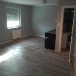 Ukończone wnętrze z szarą kolorystyką ścian i podłogą wyłożoną płytkami imitującymi drewno, widoczne okno z żaluzjami i fragment otwartego wejścia do łazienki.