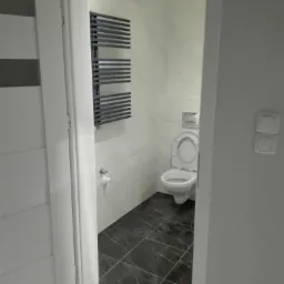 Widok z korytarza na wykończoną łazienkę z czarną marmurową podłogą, białą toaletą i grzejnikiem drabinkowym na ścianie wyłożonej białymi płytkami imitującymi marmur.