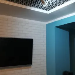 Ściana wykończona białą cegłą dekoracyjną, na niej zawieszony telewizor; fragment sufitu z podwieszaną ozdobną płytą z geometrycznym wzorem; ściana pomalowana na niebiesko.