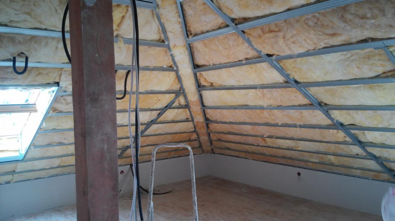 Poddasze w trakcie prac wykończeniowych: widoczne ocieplenie wełną mineralną między metalowymi profilami konstrukcji sufitu podwieszanego, drewniany słup, przewody elektryczne oraz okno dachowe.