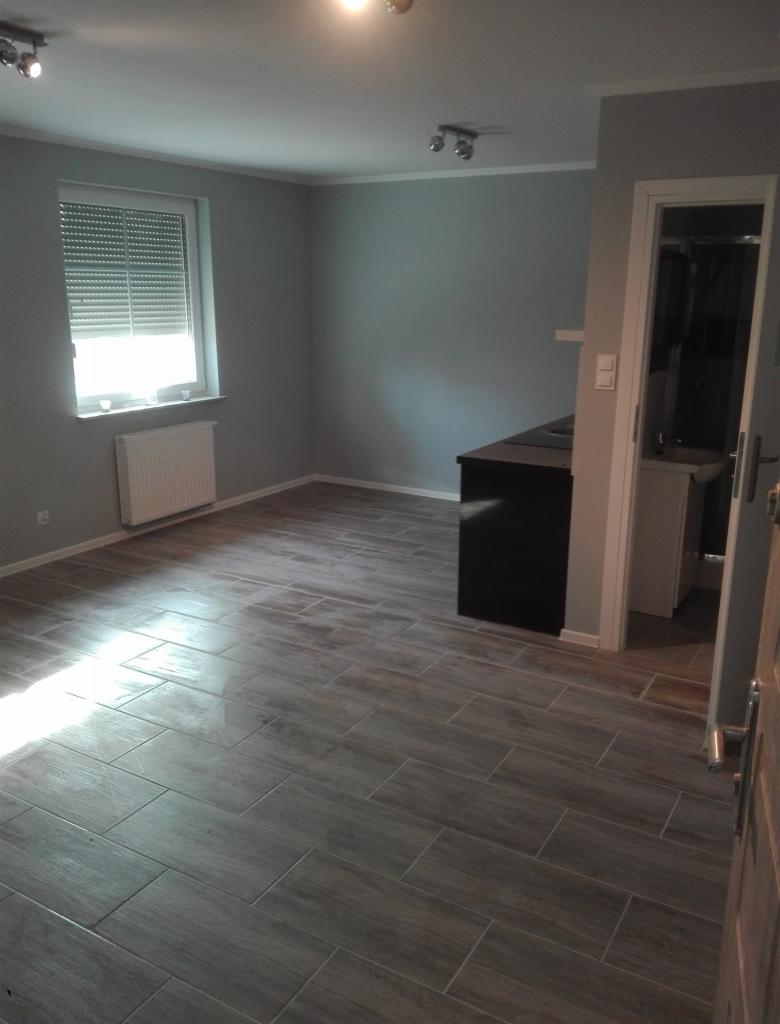 Ukończone wnętrze z szarą kolorystyką ścian i podłogą wyłożoną płytkami imitującymi drewno, widoczne okno z żaluzjami i fragment otwartego wejścia do łazienki.