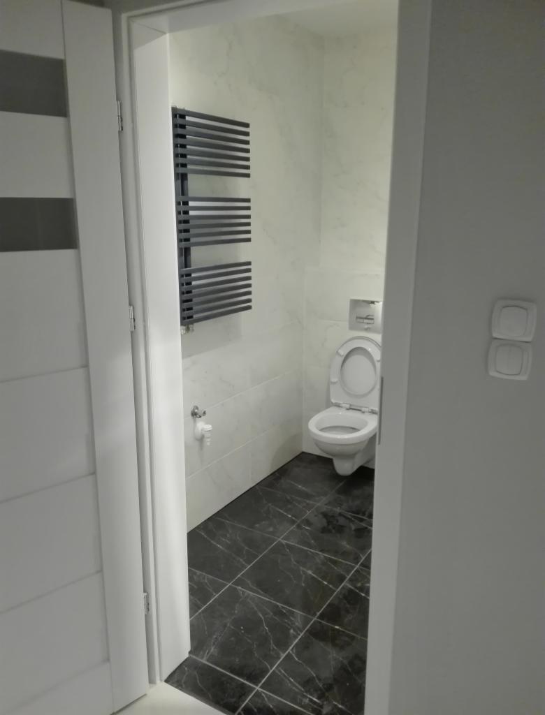 Widok z korytarza na wykończoną łazienkę z czarną marmurową podłogą, białą toaletą i grzejnikiem drabinkowym na ścianie wyłożonej białymi płytkami imitującymi marmur.