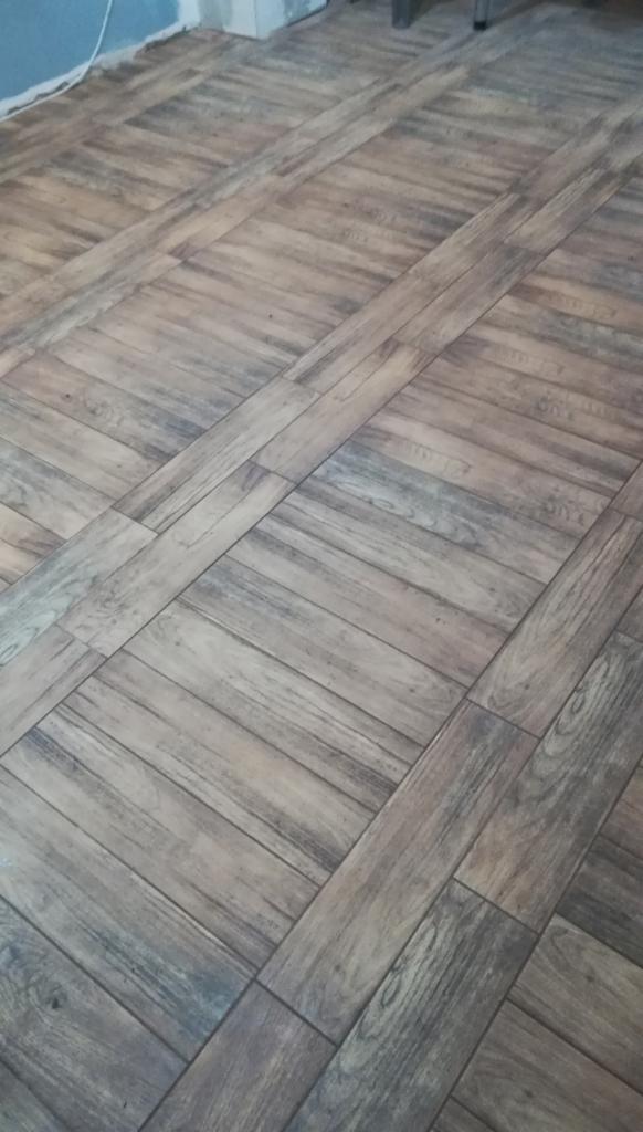 Ułożona pod kątem podłoga z płytek imitujących drewno w odcieniach brązu i szarości, z widoczną fugą i fragmentem niebieskiej ściany w tle.