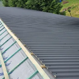 Częściowo pokryty, ciemnoszary dach z blachy trapezowej, z widoczną konstrukcją drewnianą i membraną dachową, na tle zieleni.