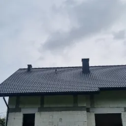 Nowy, grafitowy dach z blachodachówki na budynku w stanie surowym otwartym, widoczne dwa kominy i orynnowanie.