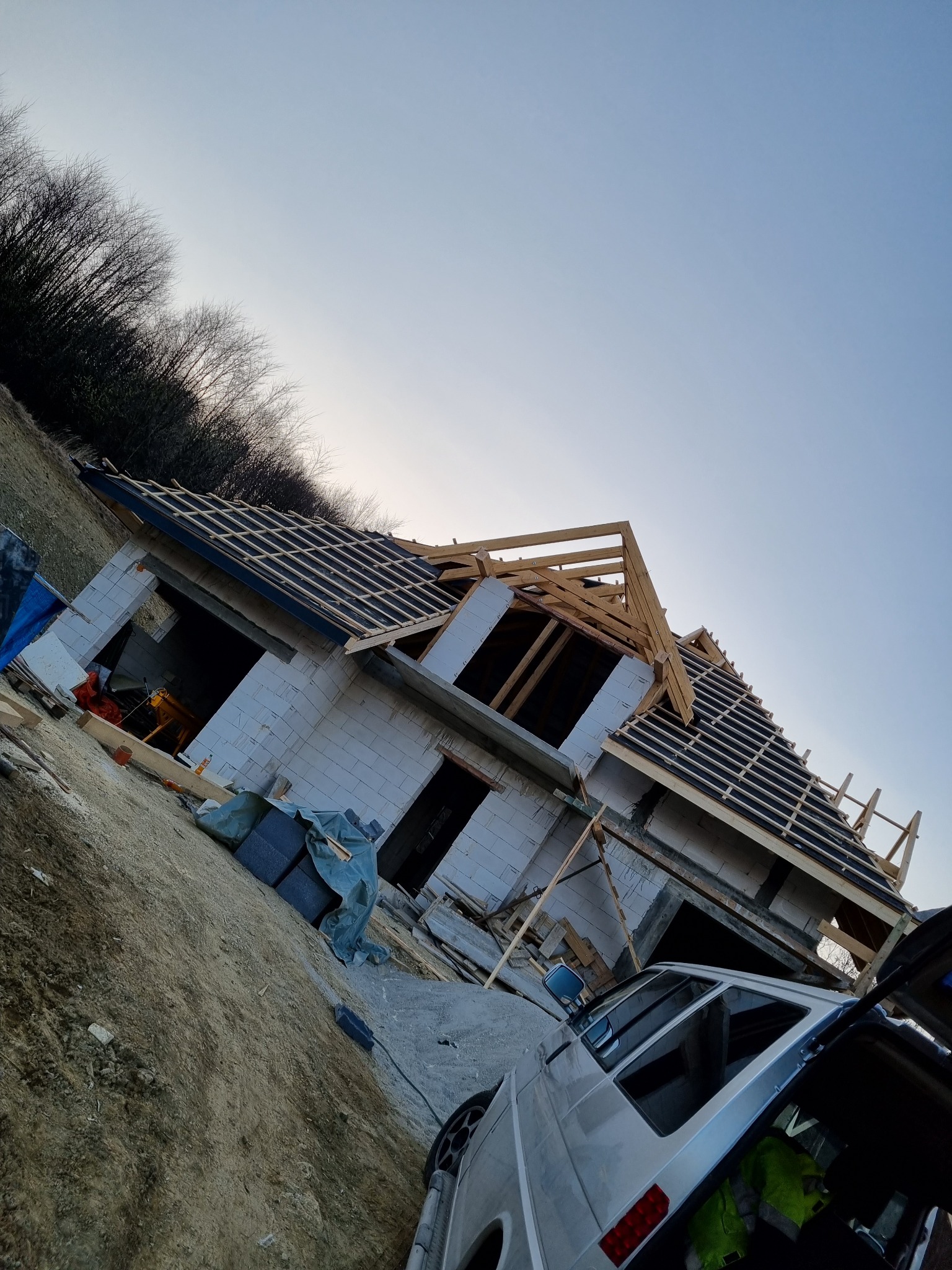 Budowa domu jednorodzinnego w trakcie: widoczne ściany z betonu komórkowego, przygotowane podłoże pod pokrycie dachu z deskami i krokwiami, samochód dostawczy z otwartym bagażnikiem na pierwszym...