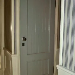 Szara, wewnętrzna, drewniana płycinowa drzwi z pionowymi żłobieniami, zamontowana w białej ościeżnicy z dekoracyjnymi listwami przypodłogowymi, na tle tapety w pionowe paski.