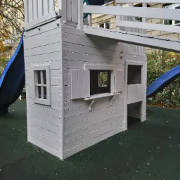 Drewniany domek zabaw dla dzieci z niebieską zjeżdżalnią, pomalowany na biało, z małymi oknami z okiennicami, ustawiony na zielonej, gumowej nawierzchni placu zabaw.