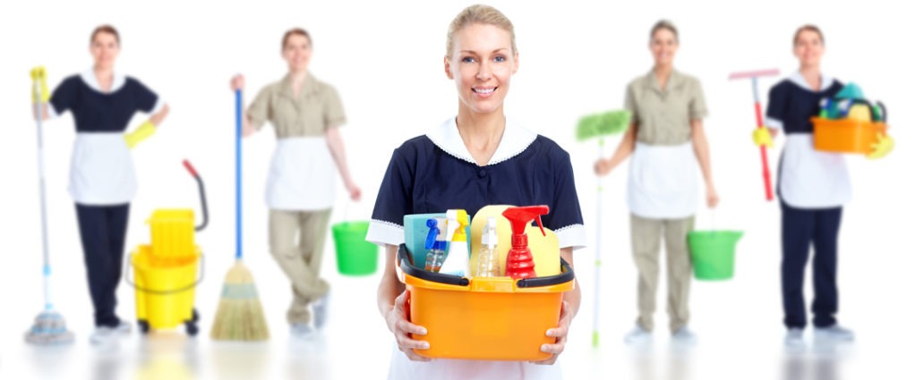 Uśmiechnięta blondynka w uniformie pokojówki prezentuje pomarańczowy pojemnik z różnymi środkami czystości, w tle widoczne inne osoby z mopami, wiadrami i szczotkami.