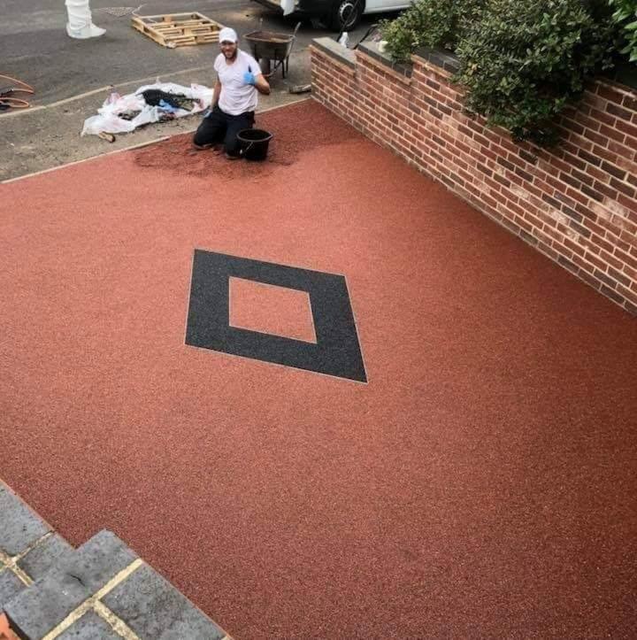 Wykończony taras kamienny z żywicy epoksydowej w kolorze ceglastym z czarnym rombem pośrodku, pracownik w białej koszulce i czarnych spodniach na kolanach podczas aplikacji żywicy.