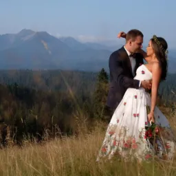 Para młoda w objęciach na tle górskiego krajobrazu. Panna młoda w sukni zdobionej folkowymi motywami kwiatowymi, pan młody w ciemnym garniturze z bordową muchą.