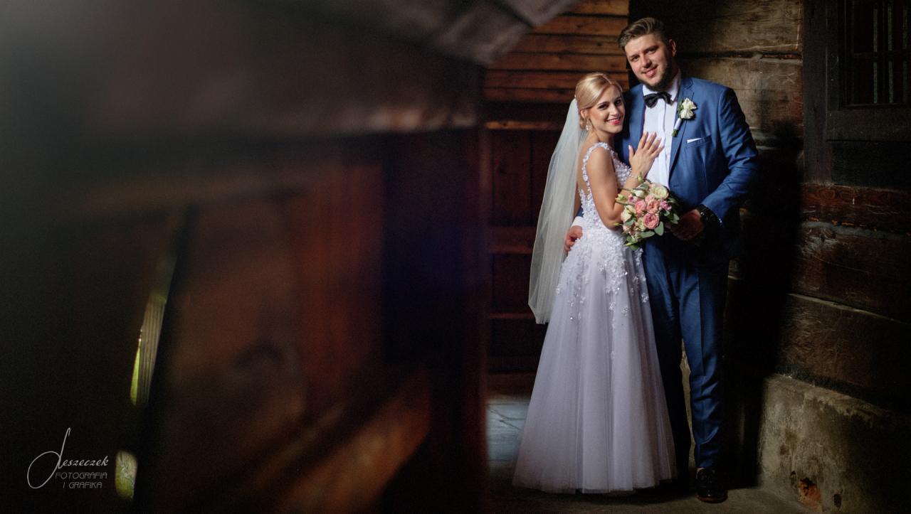 Uśmiechnięta para młoda w strojach ślubnych pozuje na tle drewnianej ściany i kamiennej podłogi, bukiet różowych kwiatów w rękach panny młodej, niebieski garnitur pana młodego.