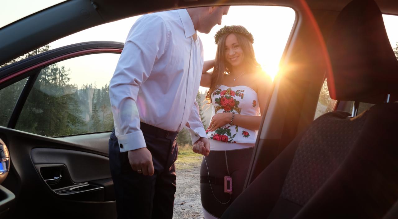 Kobieta w kwiecistej sukience i wianku na głowie, opierająca się o otwarte drzwi samochodu, mężczyzna trzyma ją za rękę, w tle las i zachodzące słońce.