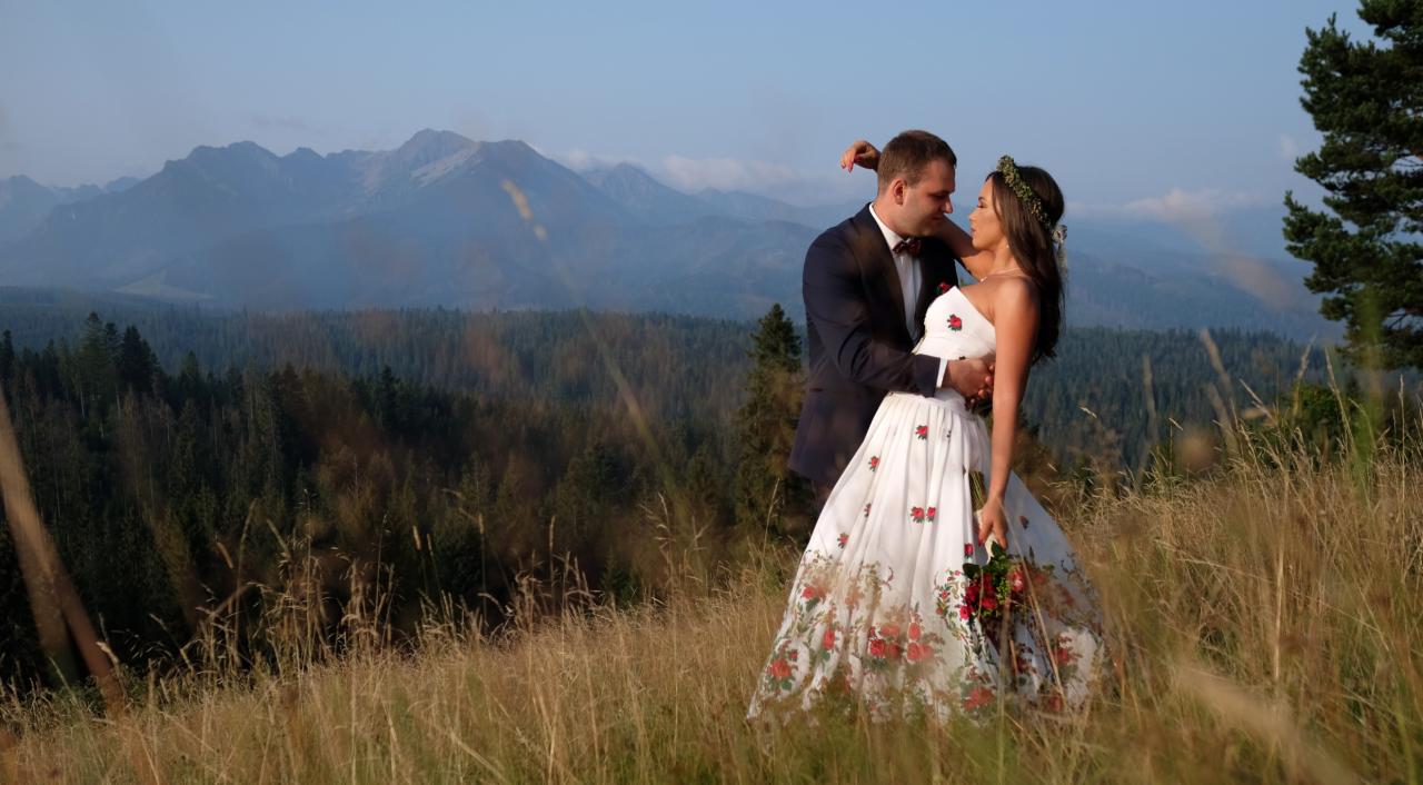 Para młoda w objęciach na tle górskiego krajobrazu. Panna młoda w sukni zdobionej folkowymi motywami kwiatowymi, pan młody w ciemnym garniturze z bordową muchą.
