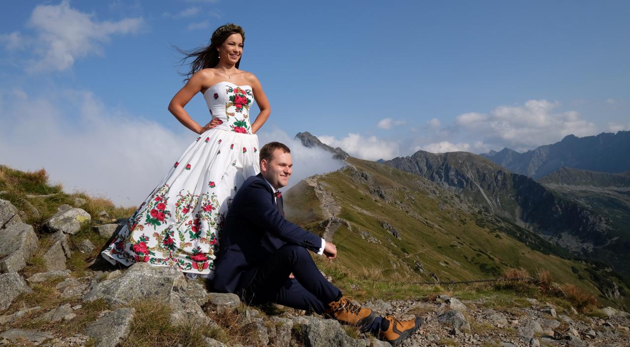 Para młoda w strojach ludowych na tle górskiego krajobrazu; panna młoda w białej sukni z czerwonymi kwiatami, pan młody w ciemnym garniturze, oboje uśmiechnięci, Tatry w tle.