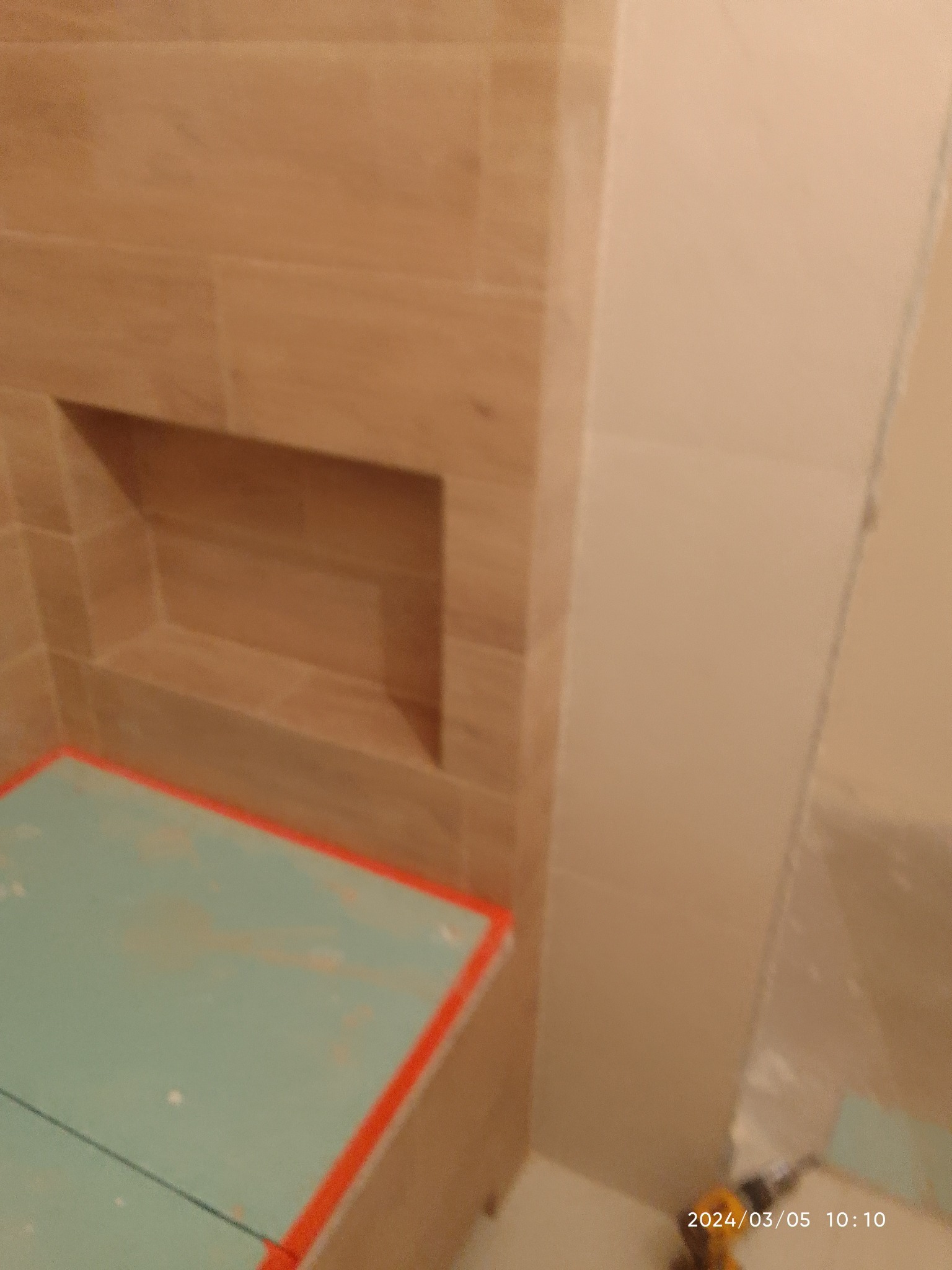 Niedokończona ściana łazienki z wnęką wyłożoną płytkami imitującymi drewno, widoczny fragment podłogi zabezpieczony płytą styropianową z czerwoną obwódką.