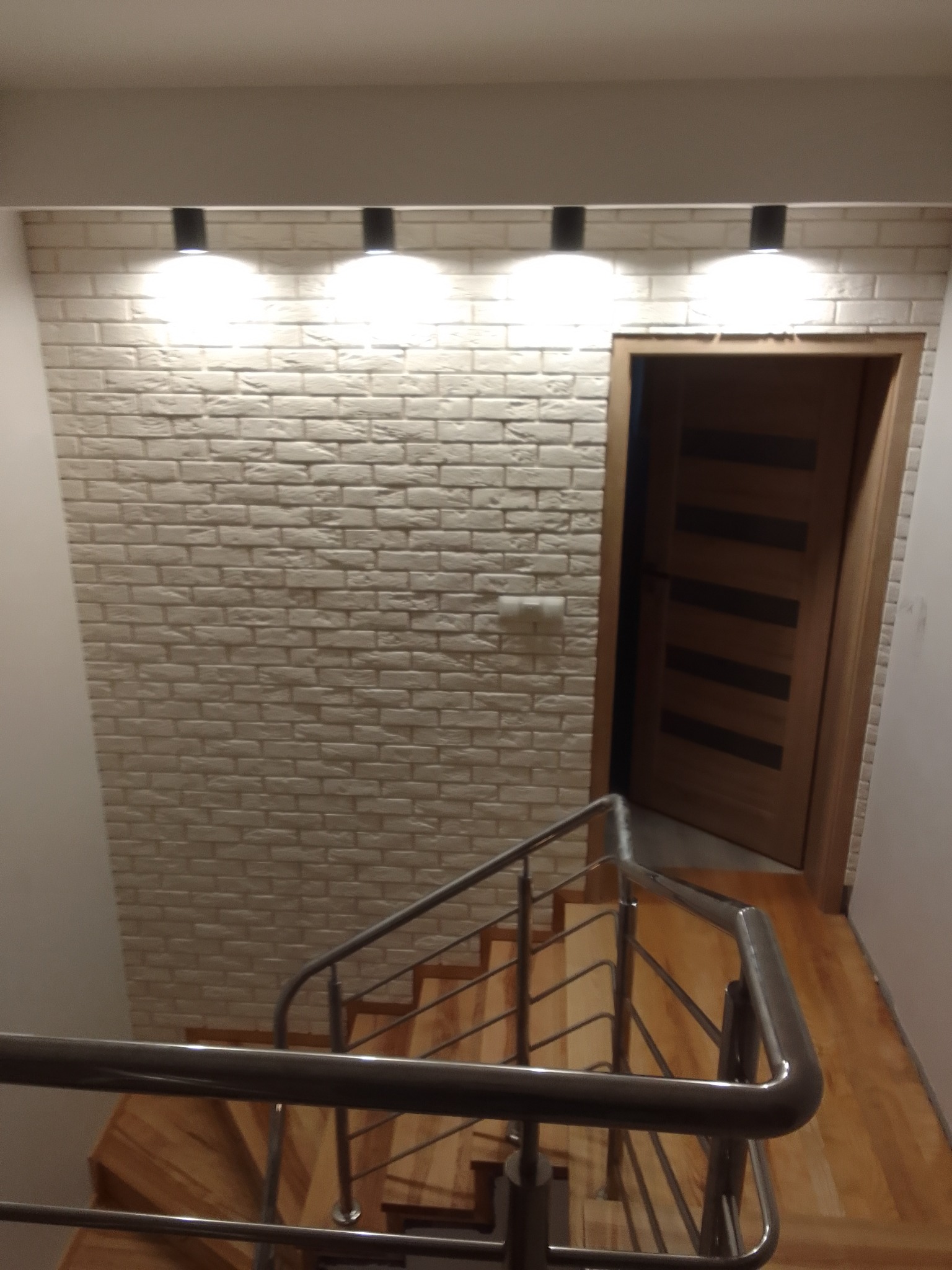 Klatka schodowa z drewnianymi schodami i metalową balustradą, ściana wyłożona cegłą dekoracyjną w odcieniach bieli, oświetlona czterema punktowymi lampami sufitowymi, widoczna drewniana ościeżnica...