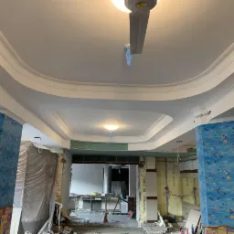 Wnętrze w trakcie remontu; widoczne kolumny z niebieską tapetą w rybki, białe sufity z dekoracyjnymi listwami, odsłonięte instalacje i resztki gruzu na podłodze.