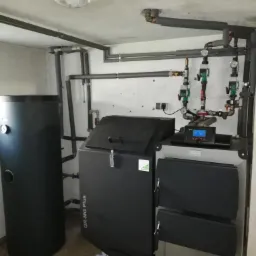 Nowoczesny piec na paliwo stałe z zasobnikiem, obok zbiornik buforowy, instalacja z zaworami i sterownikiem, widoczne rury grzewcze pod sufitem.