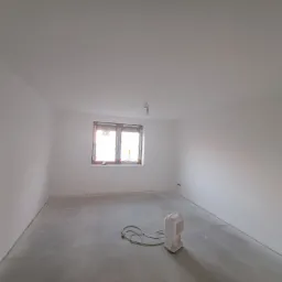 Pusty, świeżo wykończony pokój z gładkimi, białymi ścianami i sufitem; na podłodze widoczny sprzęt do osuszania w rogu pokoju.