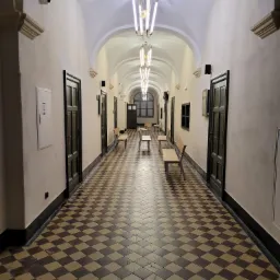 Długi korytarz z łukowatym sufitem, oświetlony nowoczesnymi lampami wiszącymi, z szachownicową posadzką z płytek w odcieniach brązu i beżu, z ciemnymi drzwiami po bokach i ławkami wzdłuż ścian.