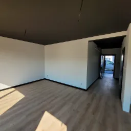 Pusty pokój po remoncie z białymi ścianami, ciemnym sufitem i panelami podłogowymi, widoczne gniazdka elektryczne i cień padający na podłogę.