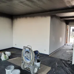 Wnętrze w trakcie malowania natryskowego, widoczne ściany i sufit pokryte szarą farbą, zabezpieczona folią podłoga, agregat malarski na wózku z nawiniętym wężem, dwa białe wiadra z farbą...