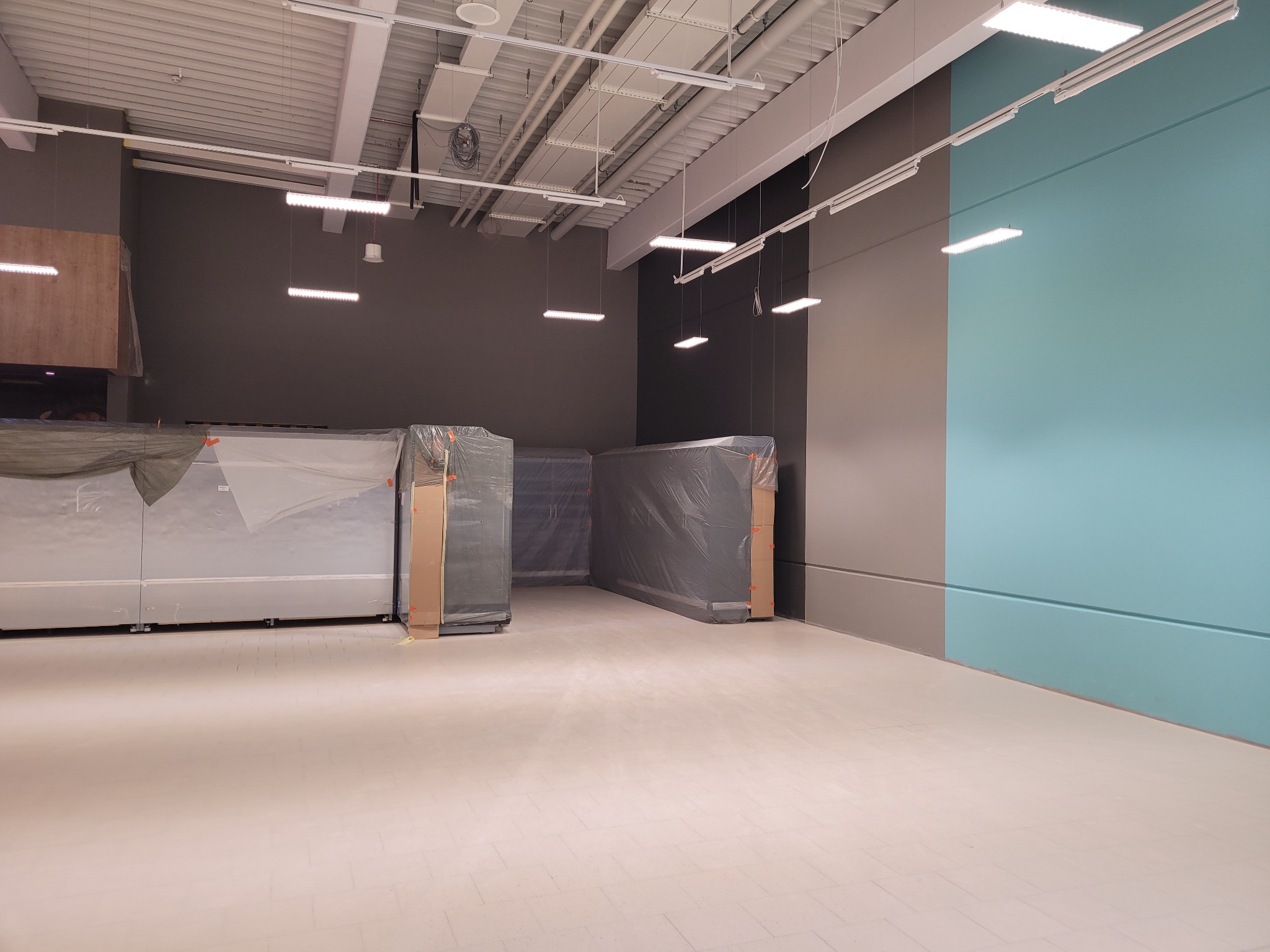 Pusta przestrzeń biurowa przed umeblowaniem, świeżo po malowaniu ścian na szaro i turkusowo, z podłogą w jasnym odcieniu, z meblami w folii ochronnej.