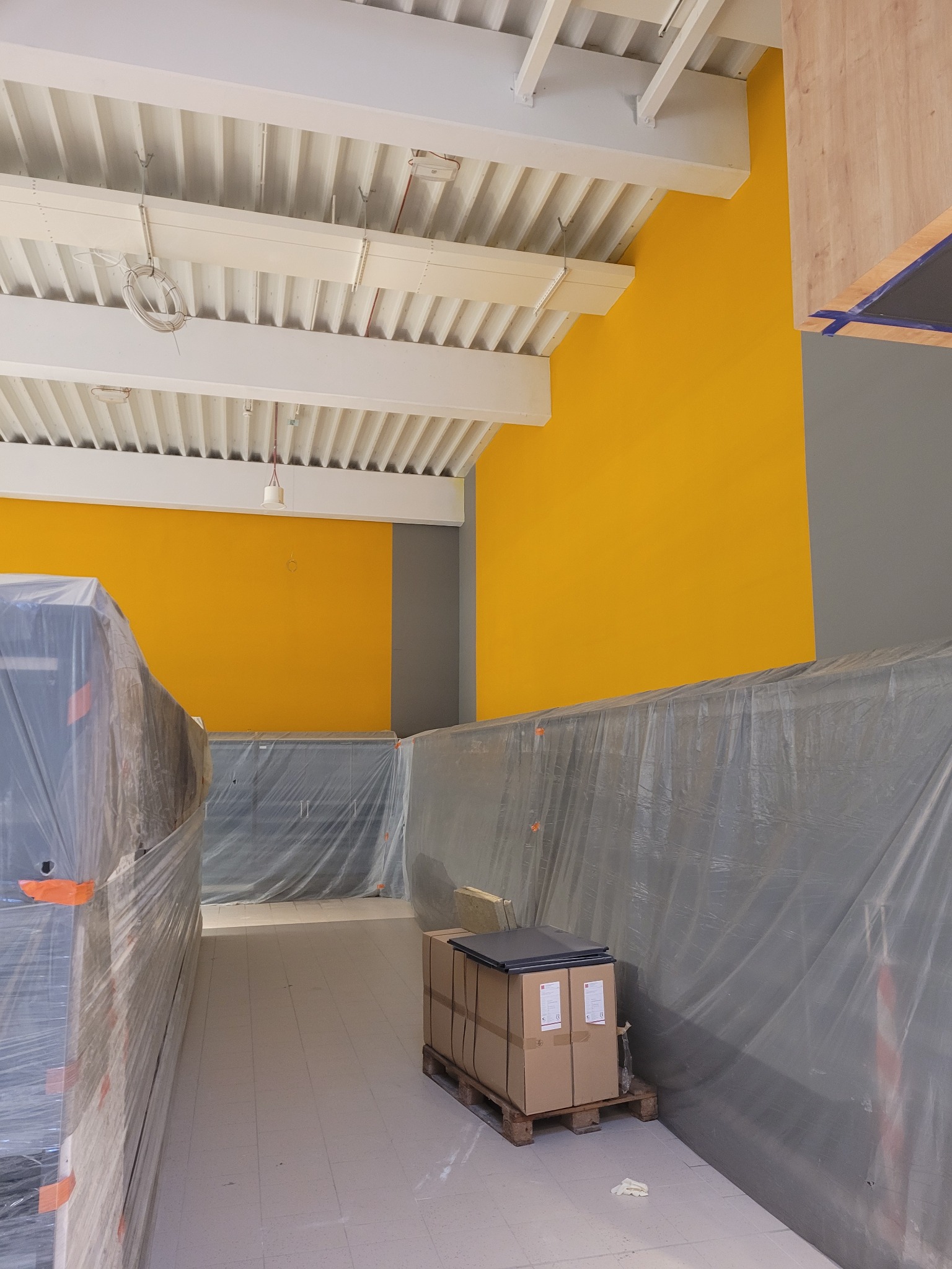Wnętrze biura w trakcie przygotowań do malowania, ściany pomalowane na żółto i szaro, meble zabezpieczone folią, paczki na palecie, widoczny surowy sufit z instalacjami.