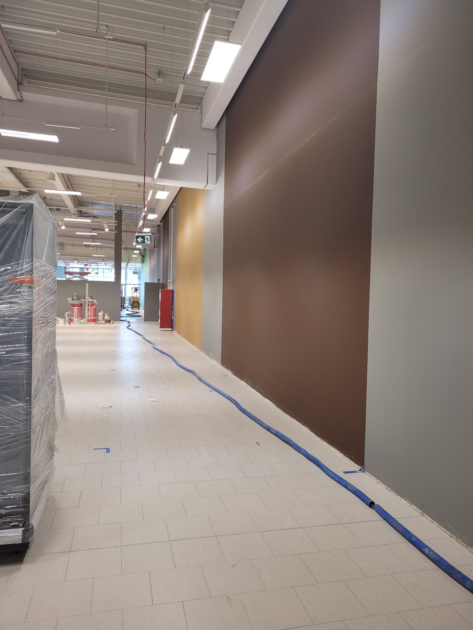 Wnętrze sklepu w trakcie odświeżania, ściany pomalowane na brązowo, szaro i żółto, niebieski wąż zabezpieczający podłogę, w tle widoczne materiały budowlane i rusztowanie.